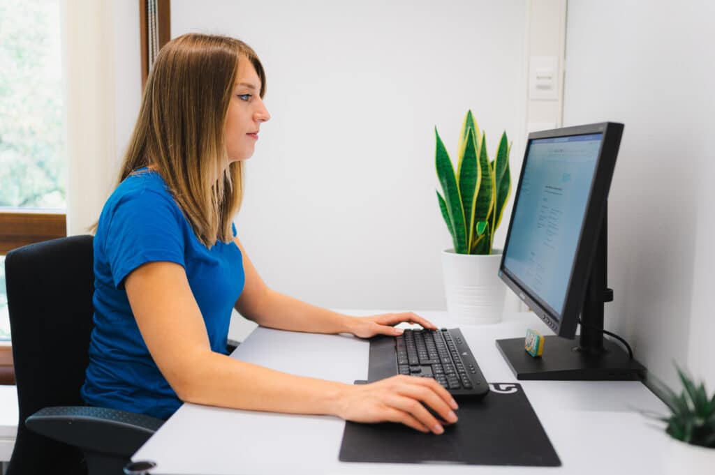 Céline Matter in blauem Oberteil arbeitet an einem Schreibtisch in einem hellen Büro, vor ihr ein Computerbildschirm, eine Tastatur und eine Pflanze.