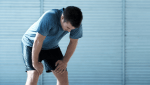 Ein erschöpfter Sportler in blauem T-Shirt und schwarzer Shorts beugt sich nach einer Trainingseinheit vor und stützt sich auf seine Knie, während er versucht, Atem zu holen.