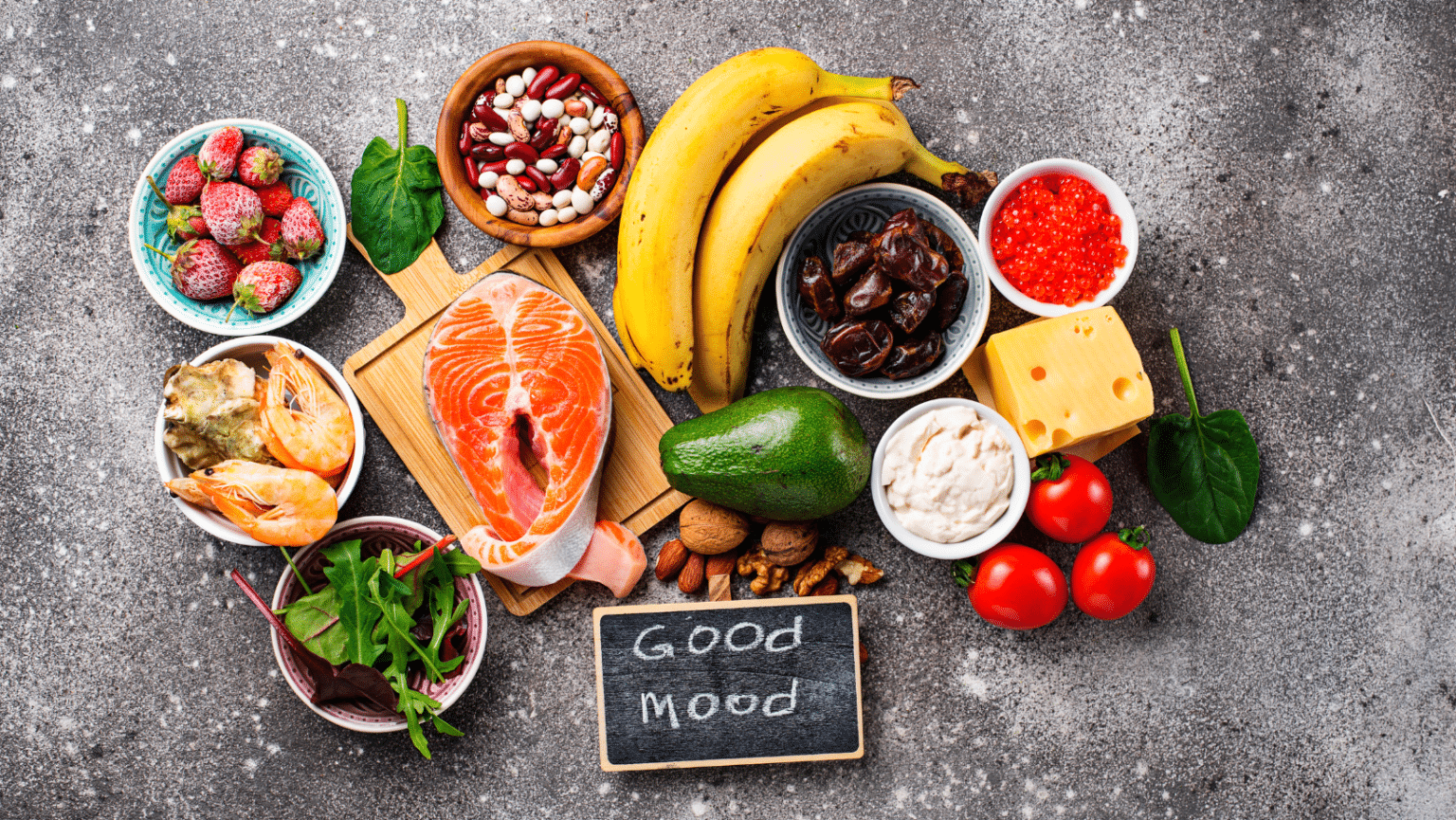 Eine Vielzahl von nährstoffreichen Lebensmitteln angeordnet um ein Schild mit der Aufschrift 'Good Mood', darunter Lachs, Avocado, Käse, Bananen, Beeren, Nüsse und Gemüse auf einer grauen Oberfläche